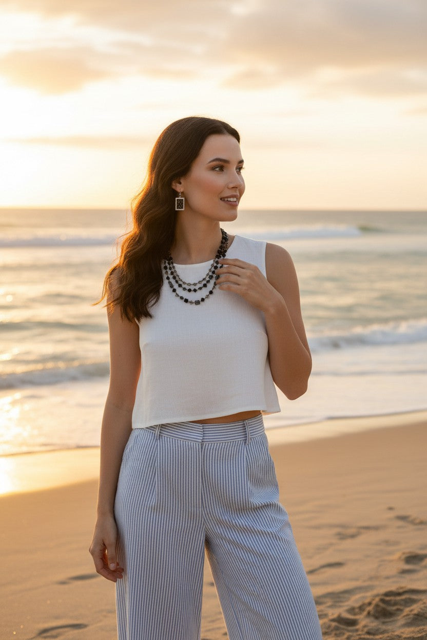 A woman stands on a sandy beach at sunset wearing a white sleeveless crop top, light blue and white pinstriped wide-leg pants, and a layered black beaded necklace.