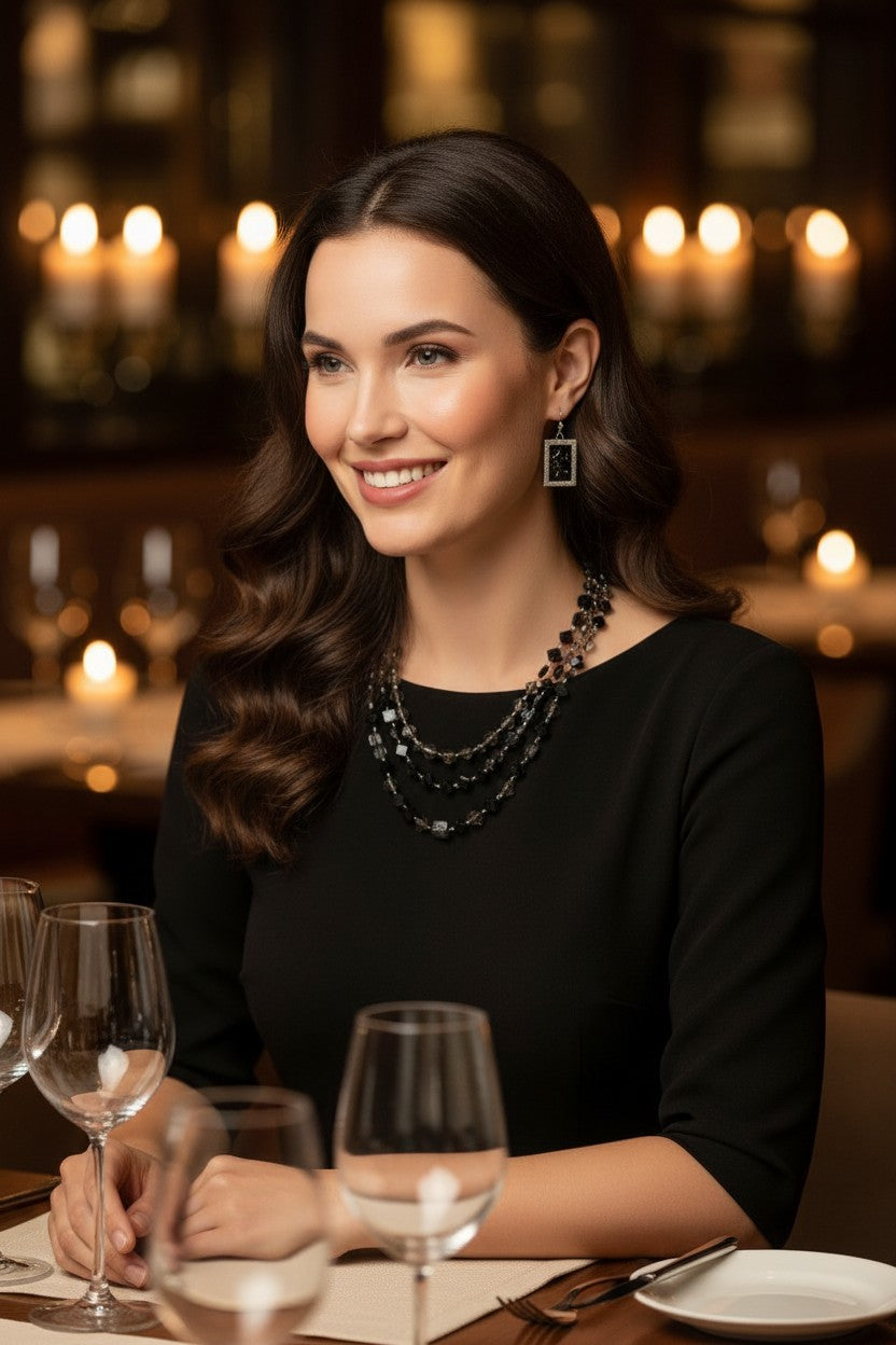 A woman with long dark brown hair sits at a restaurant table wearing a black top and layered black beaded necklaces, with wine glasses and plates in the foreground and warm candlelit background.
