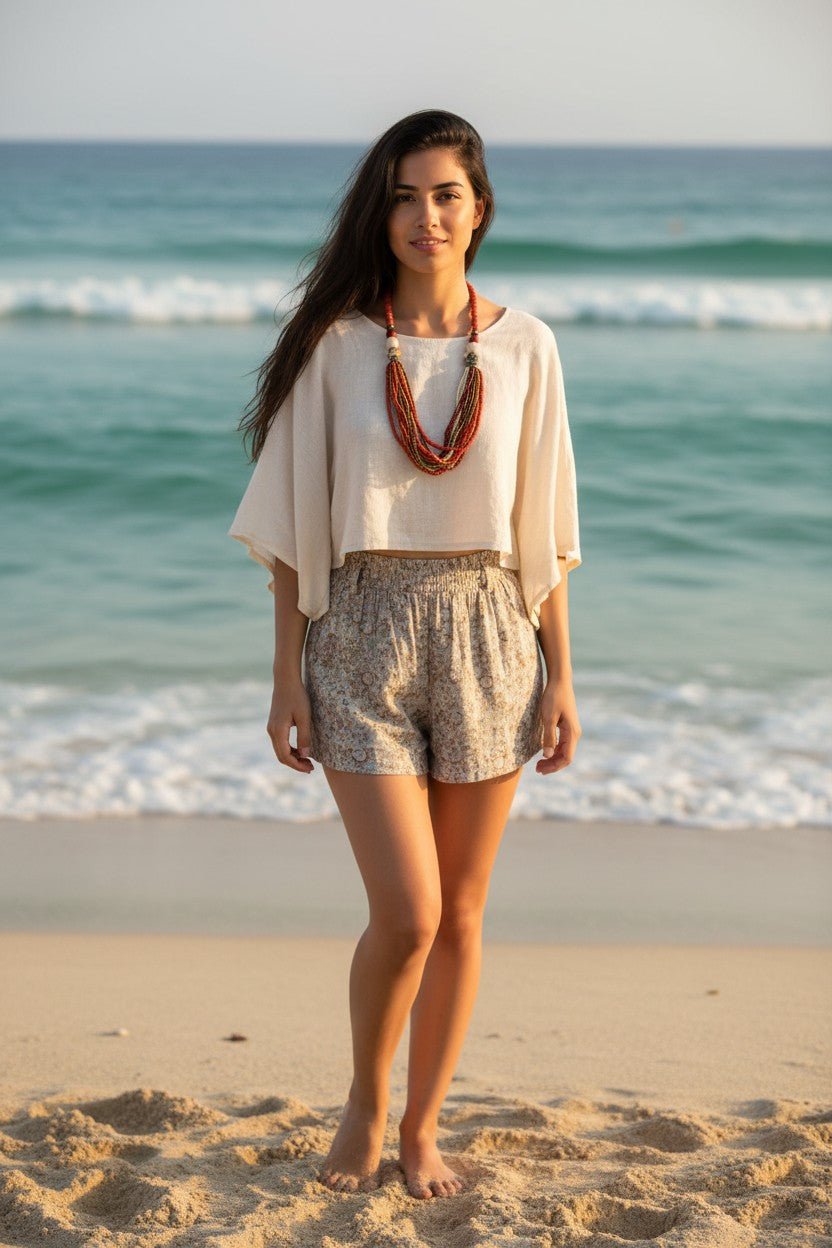 A woman stands on a sandy beach in front of the ocean waves, wearing a cream-colored short-sleeve top, patterned beige and white shorts, and a multi-strand orange necklace.