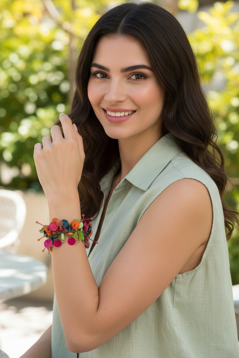 A woman with long dark hair wearing a light sage green sleeveless top and a colorful beaded bracelet, posing outdoors with a blurred green trees background.