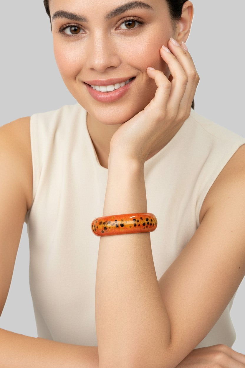 A close-up view of a smiling model wearing a high-shine, glossy orange lacquered wood bangle bracelet with a black speckled pattern.