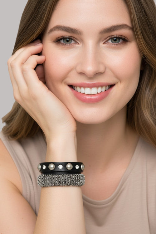 Close-up view of a model wearing a black leather strap wrap bracelet adorned with pearls, crystal studs, and a textured metallic mesh chain band.