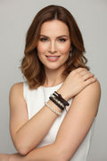 A woman with shoulder-length brown hair wearing a white sleeveless top and multiple stacked bracelets on her left wrist, posed against a light gray studio background.