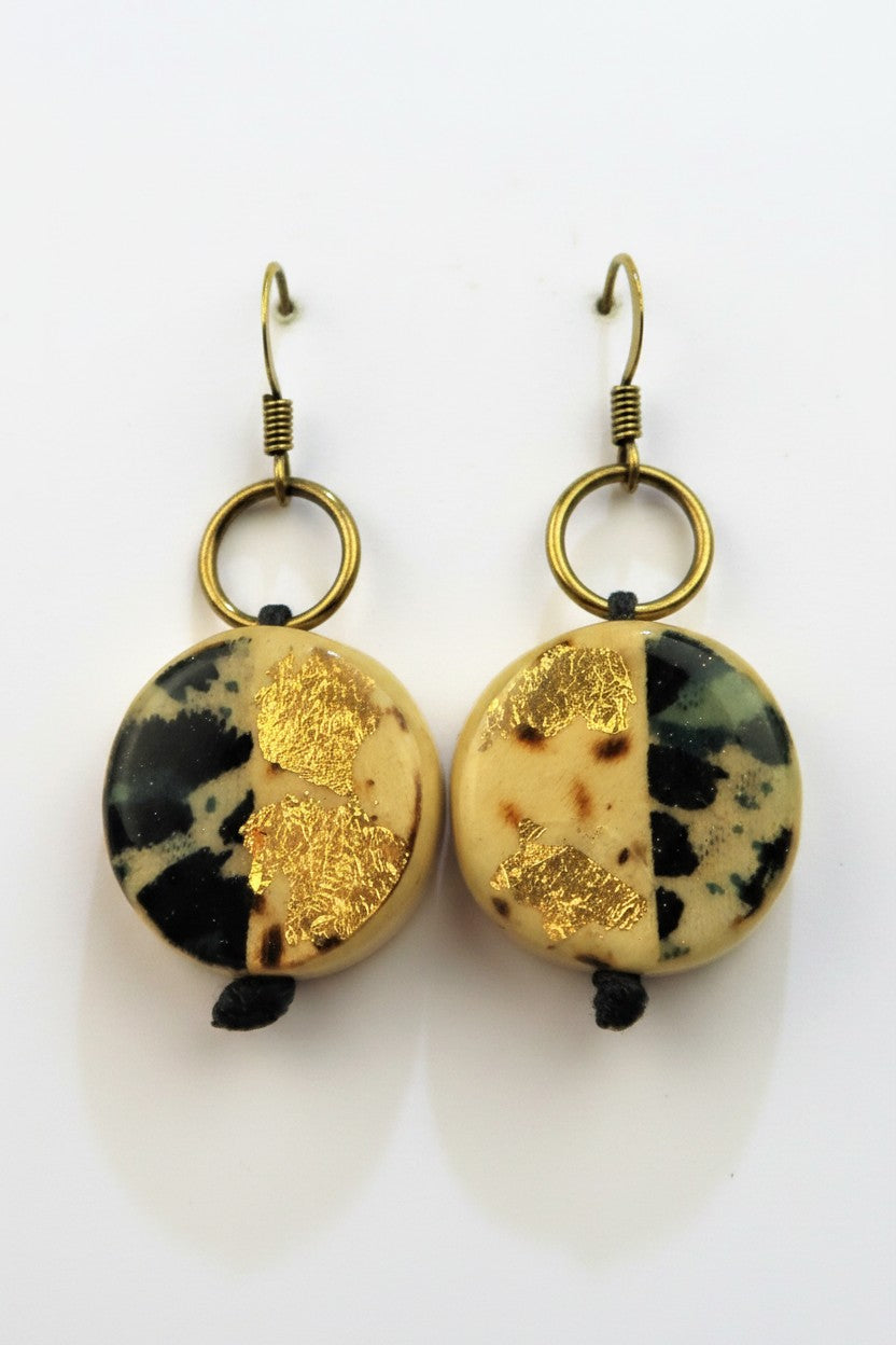 A pair of handmade dangle earrings featuring round, split-face stone beads with one half showing black and beige marbling and the other half decorated with textured gold foil. Each bead hangs from a gold-toned metal ring and a classic fishhook ear wire, set against a plain white background.