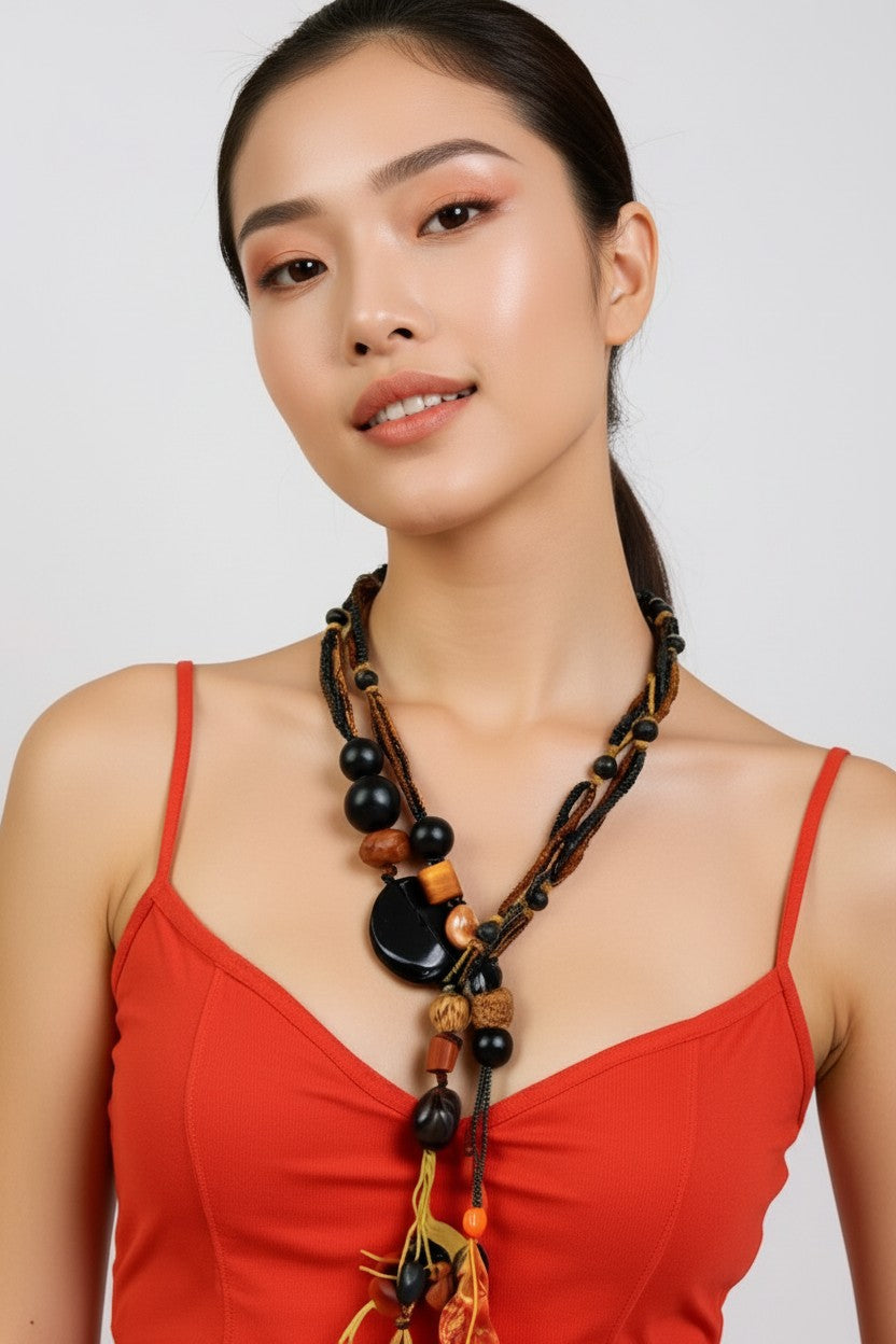 A woman wearing a bohemian-style statement necklace with dark beads, wooden elements, and tassels, paired with a red spaghetti-strap top against a light gray background.