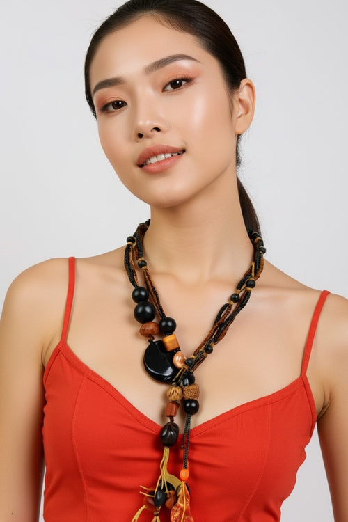 A woman wearing a bohemian-style statement necklace with dark beads, wooden elements, and tassels, paired with a red spaghetti-strap top against a light gray background.