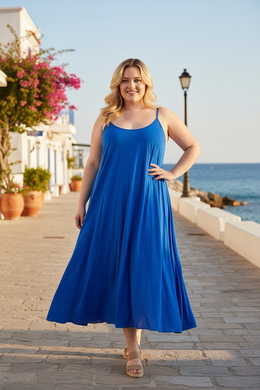 A woman modeling a vibrant royal blue sleeveless maxi dress on a sunny stone walkway by the sea, with white buildings and pink bougainvillea flowers in the background.