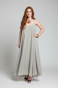 A woman modeling a sleeveless, floor-length A-line dress in a flax (light beige) color, styled with a gold necklace and bracelets, against a neutral gray studio background.