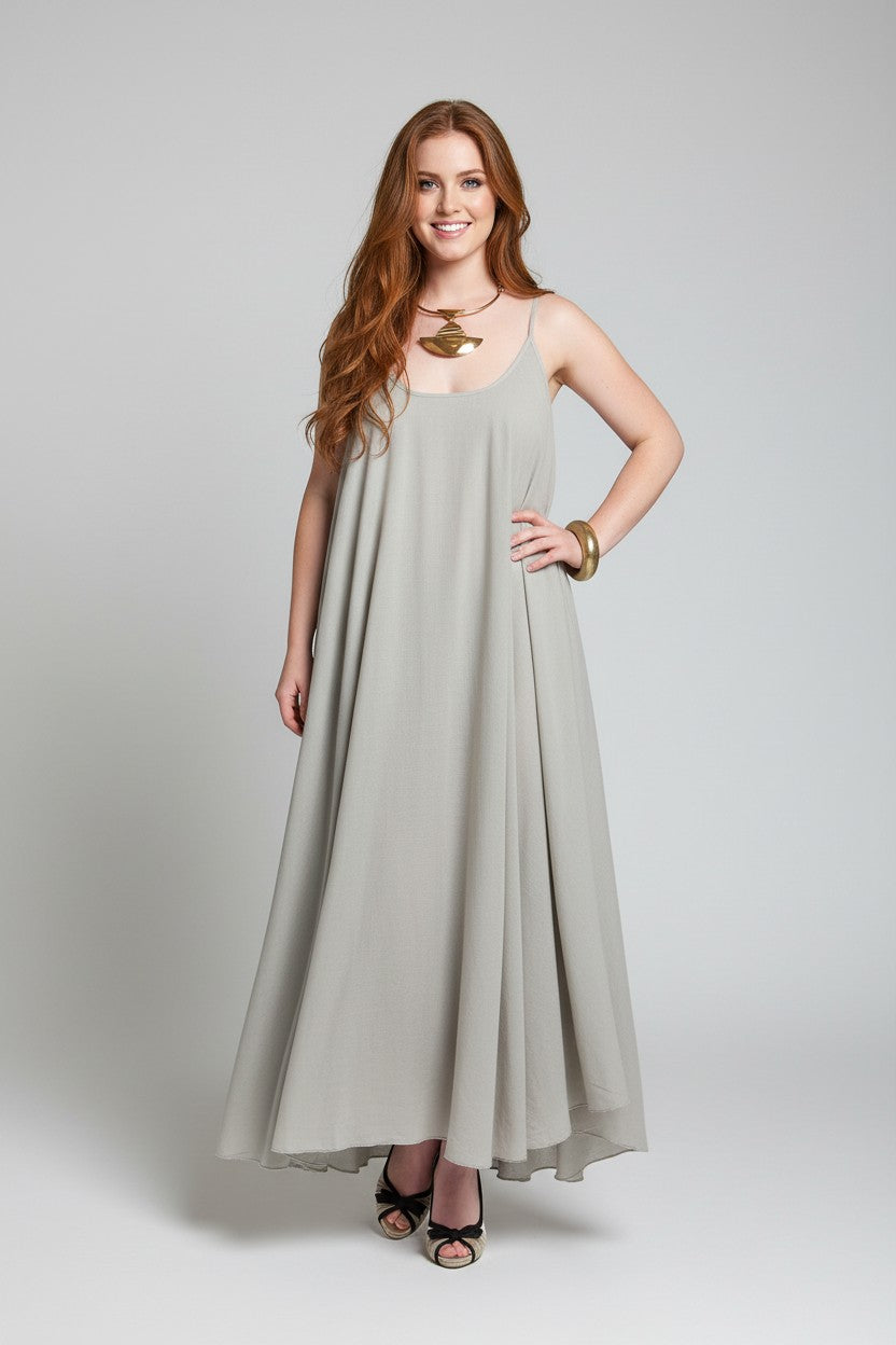 A woman modeling a sleeveless, floor-length A-line dress in a flax (light beige) color, styled with a gold necklace and bracelets, against a neutral gray studio background.