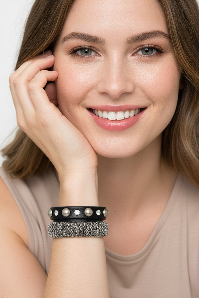 Close-up view of a model wearing a black leather strap wrap bracelet adorned with pearls, crystal studs, and a textured metallic mesh chain band.