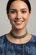 A smiling woman wearing a colorful beaded choker necklace with turquoise blue beads and multicolored accents, paired with a blue patterned top against a neutral gray background.