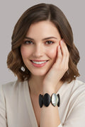 A woman with brown shoulder-length hair wearing a cream-colored top and a black and iridescent shell bracelet, posing with her hand on her cheek against a solid gray background.