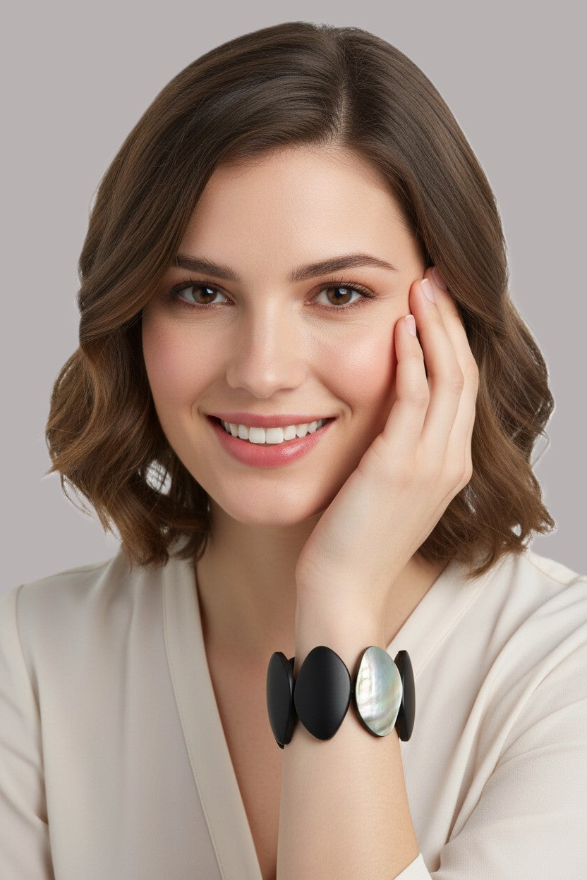 A woman with brown shoulder-length hair wearing a cream-colored top and a black and iridescent shell bracelet, posing with her hand on her cheek against a solid gray background.