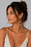 A woman with her hair in a bun is shown in profile wearing a long, dangling statement earring. The earring features a silver-toned metal setting with a round reddish-brown bead and a silver-toned cylindrical charm.