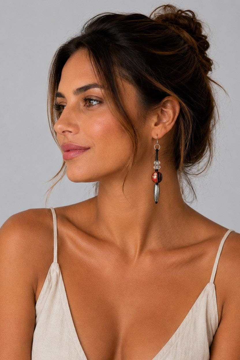 A woman with her hair in a bun is shown in profile wearing a long, dangling statement earring. The earring features a silver-toned metal setting with a round reddish-brown bead and a silver-toned cylindrical charm.
