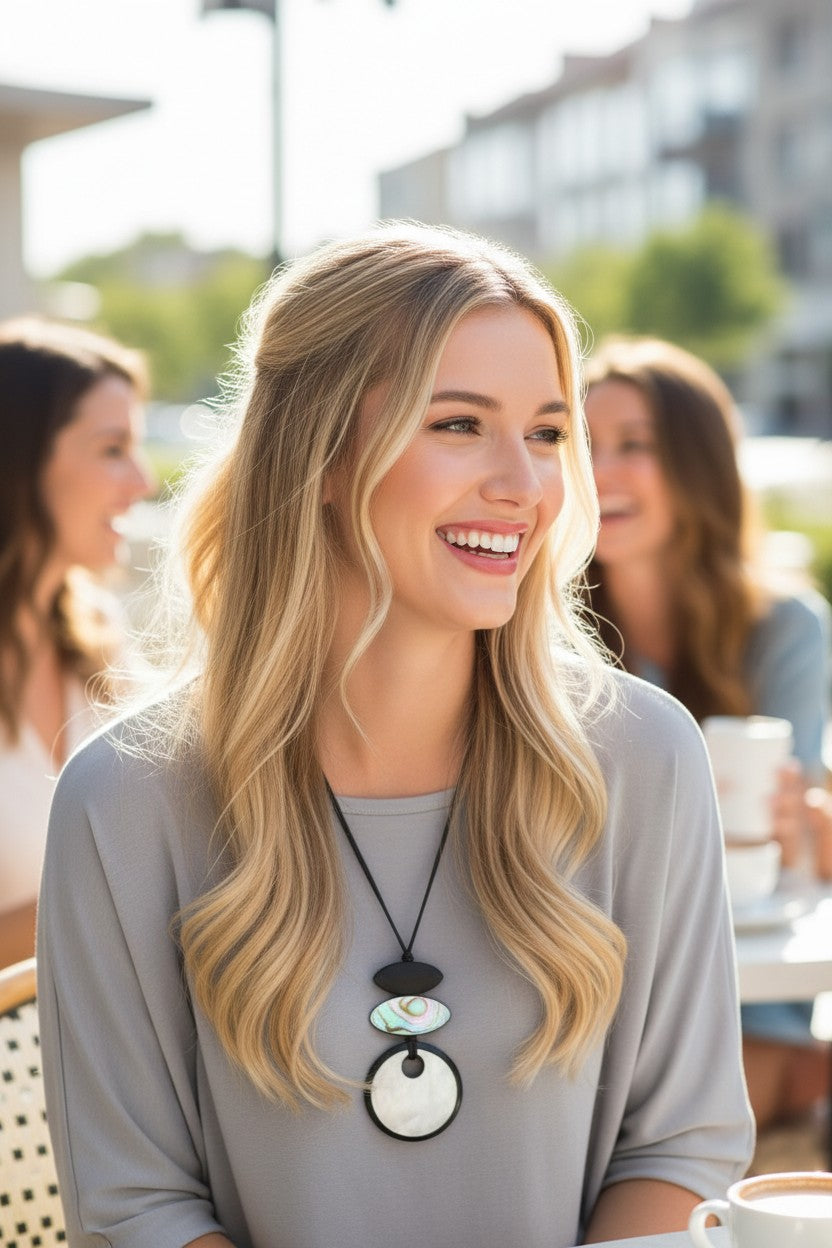 A smiling blonde woman in a gray top wears a black pendant necklace with stacked circular charms, photographed at an outdoor cafe with blurred people in the background.
