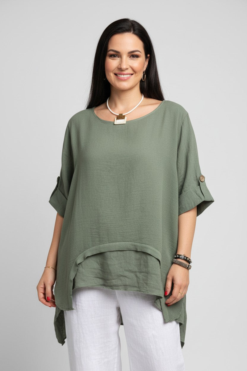 A woman modeling a loose, short-sleeved sage green top with a draped hem detail, paired with white pants, against a solid light gray background.