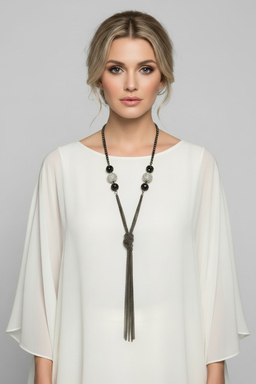 Model wearing a long gunmetal multi-chain necklace featuring a knotted tassel design, textured wire loop accents, and large silver beads with sparkling stone inlays over a white silk top.