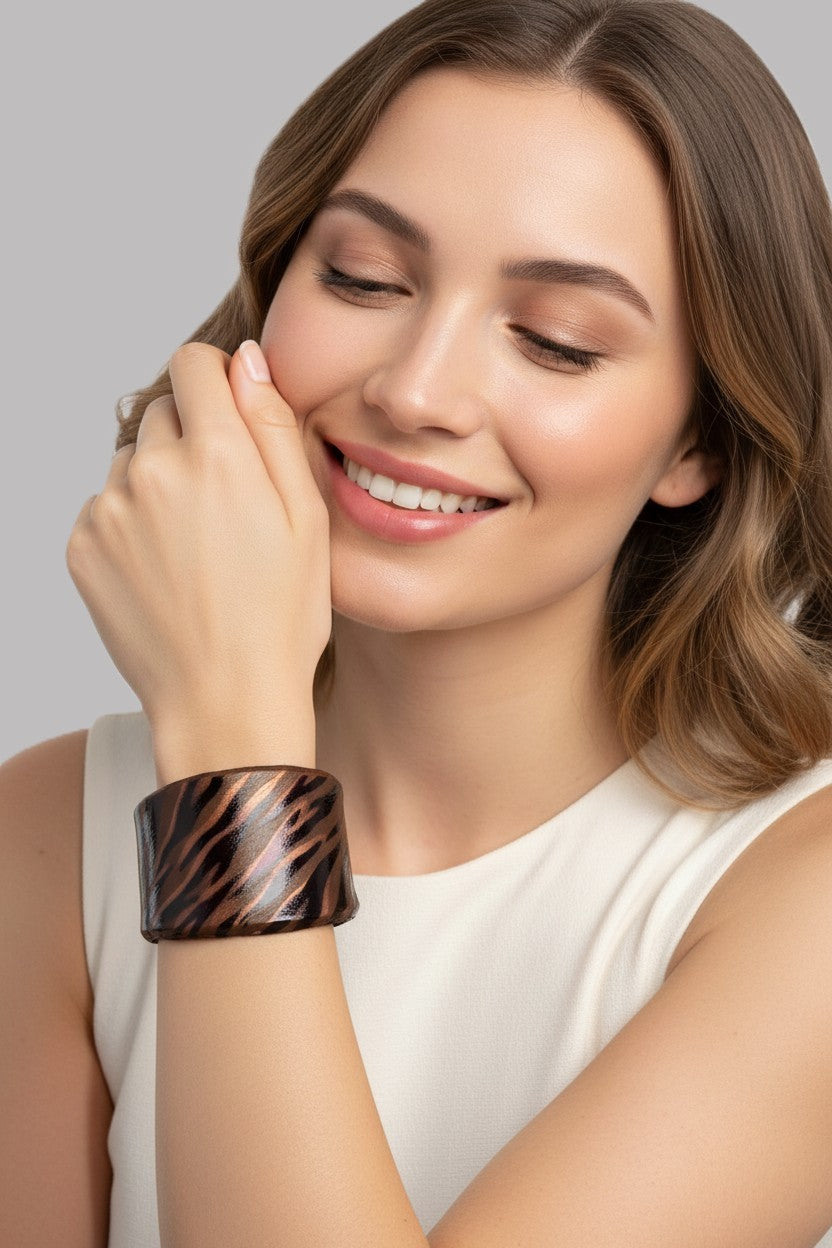 A close-up view of a smiling model wearing a wide structured leather cuff bracelet featuring a shimmering metallic bronze and dark brown animal print pattern.