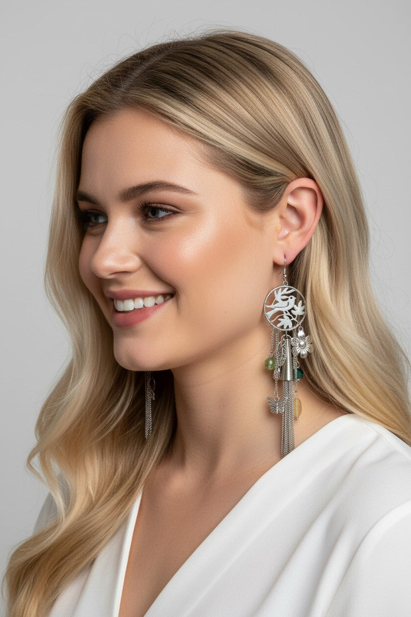 Close-up of a smiling blonde model wearing a long silver drop earring featuring a circular cutout pendant with a bird and tree design, cascading fine chains, metal charms, and green crystal beads.