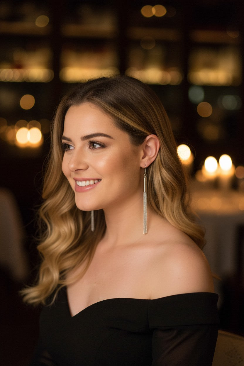 A woman with wavy blonde-highlighted hair wearing long, thin silver dangle earrings and a black off-the-shoulder top, photographed in a warmly lit indoor setting with blurred lights in the background.