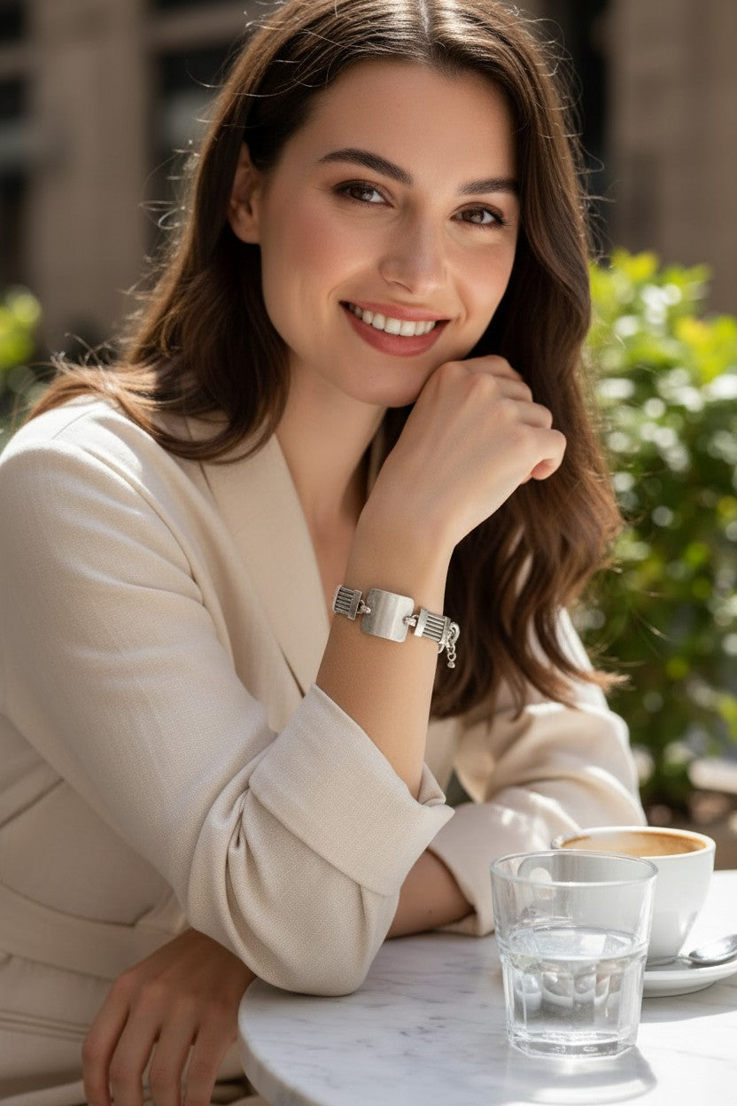 A woman with long brown hair smiles while leaning on a table at an outdoor cafe, wearing a silver rectangular analog watch with a metal link bracelet. A glass of water and a cup of coffee are on the table in front of her.